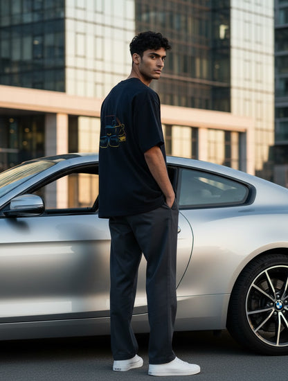 Man wearing a Print 999 black oversized graphic t-shirt with a neon car print, standing by a silver sports car.
