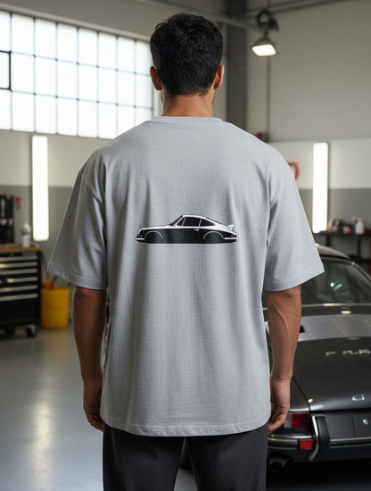 Print 999 grey oversized t-shirt with a vintage car graphic on the back, worn by a man in a garage setting.