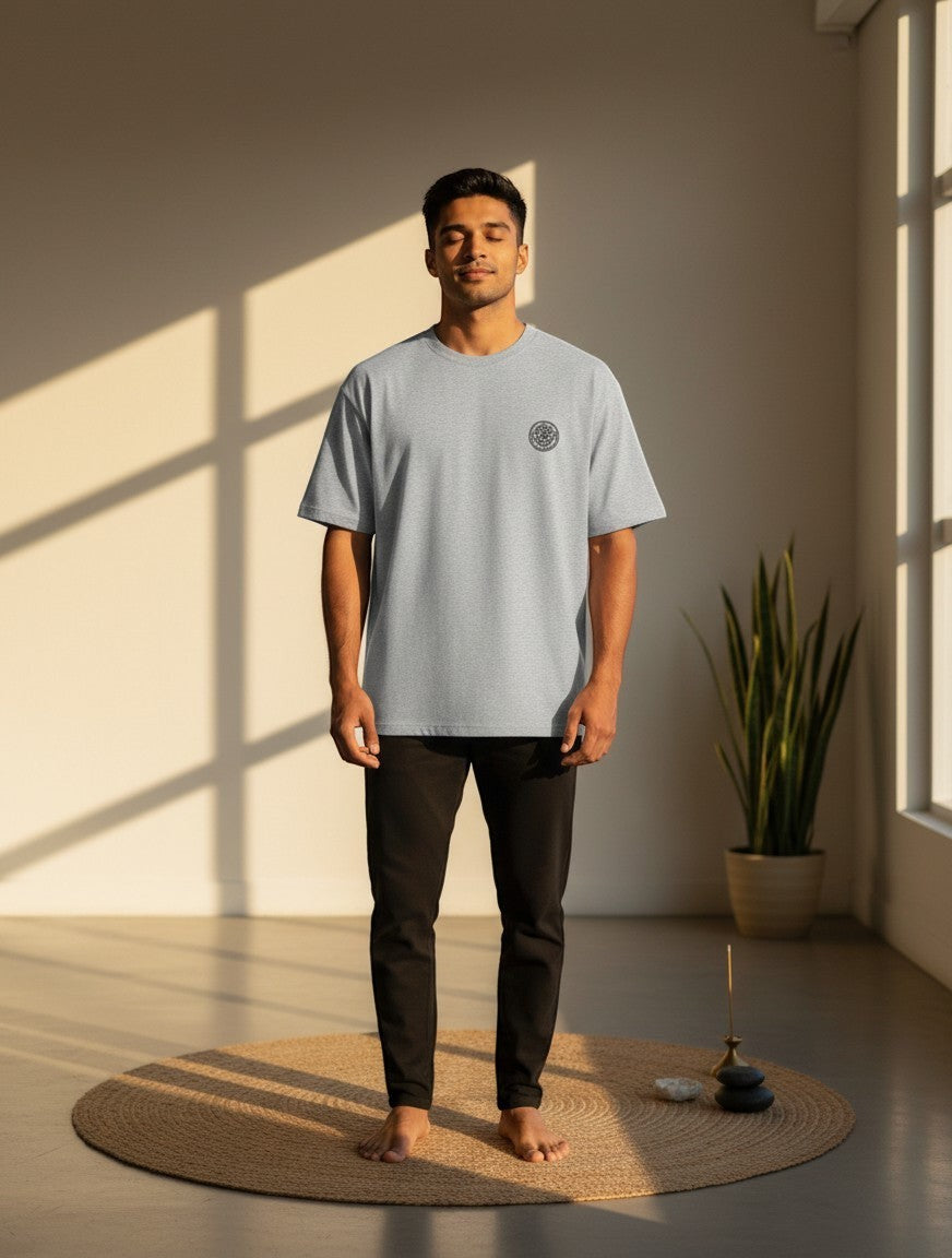 Print 999 grey oversized t-shirt with embroidered Om Sri Yantra emblem worn by a man in a sunlit room.