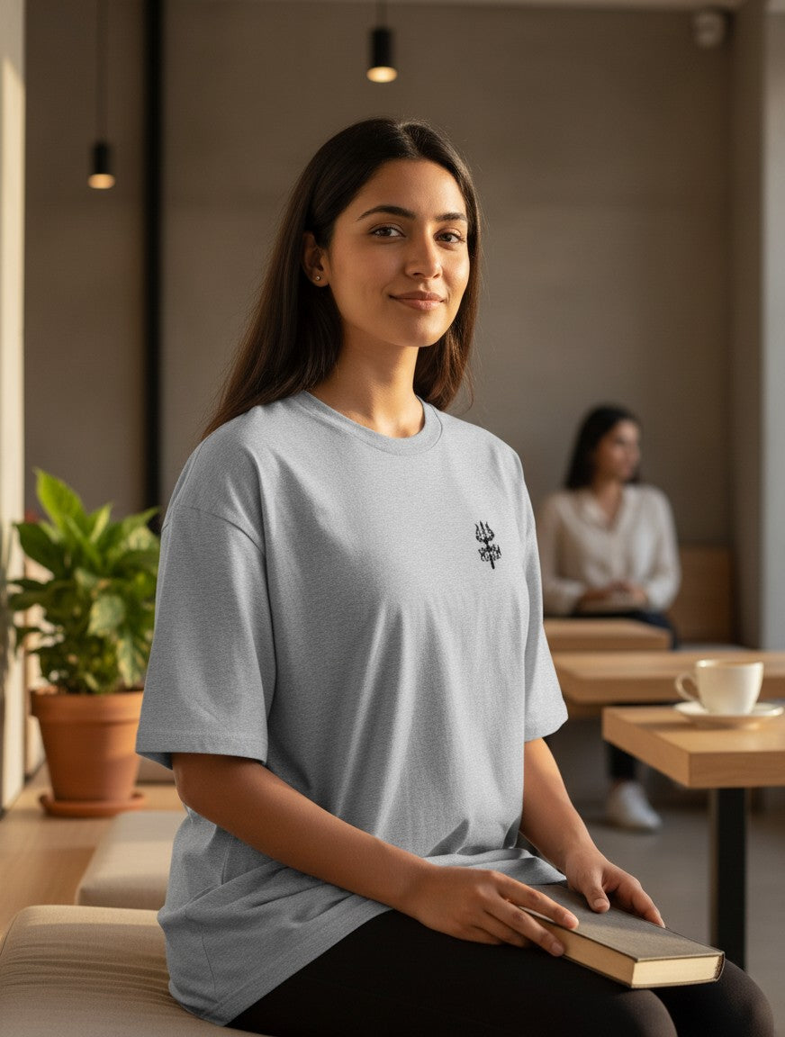 Print 999 grey oversized cotton t-shirt with embroidered Trishul emblem worn by a woman in a cafe setting.
