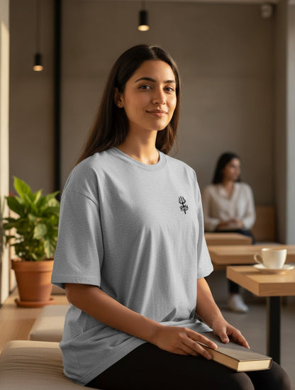 Print 999 grey oversized cotton t-shirt with embroidered Trishul emblem worn by a woman in a cafe setting.