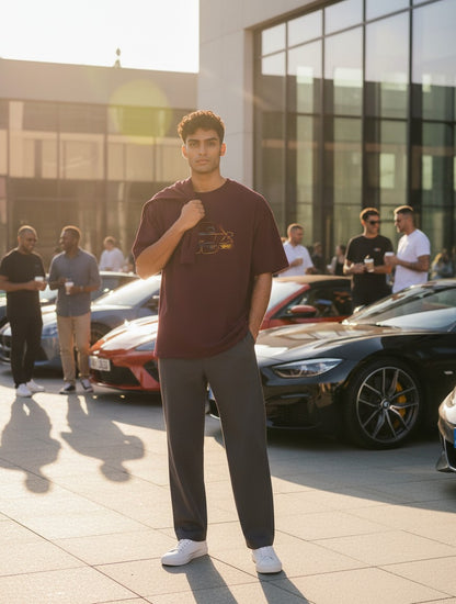 A man wearing a Print 999 maroon oversized cotton t-shirt with a graphic print, standing outdoors near luxury cars.