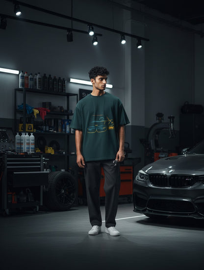 Print 999 oversized dark green cotton t-shirt with neon car graphic, worn by a man in a garage setting.
