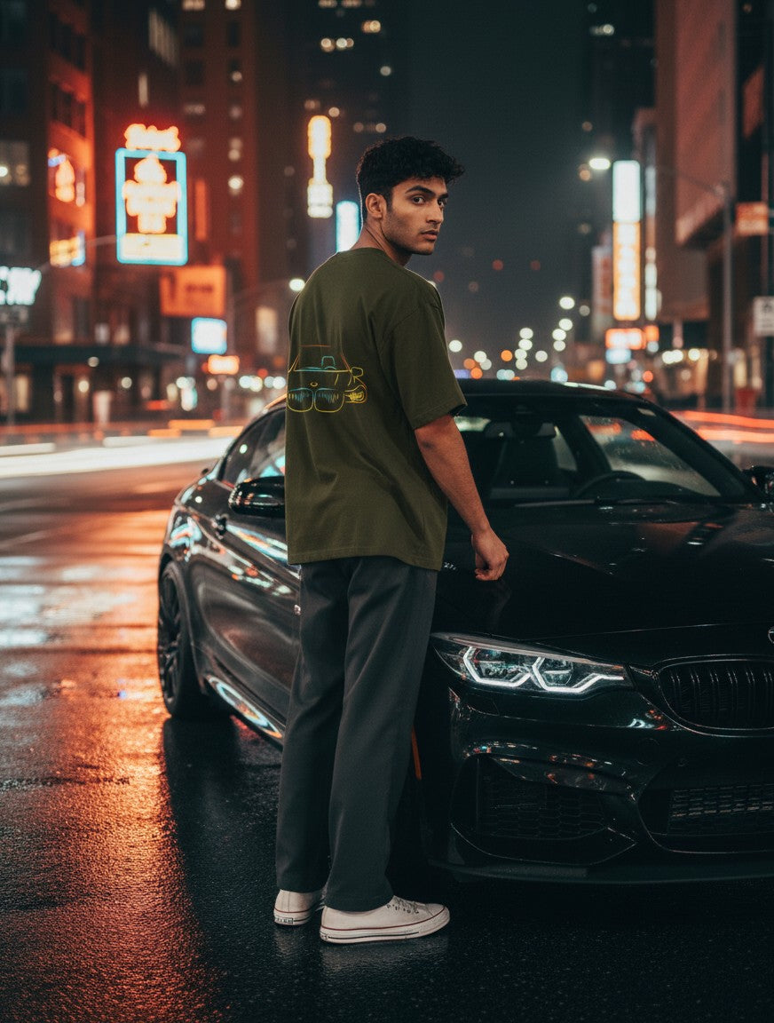 A man wearing a Print 999 olive green oversized cotton t-shirt with a neon car graphic, standing by a car at night.