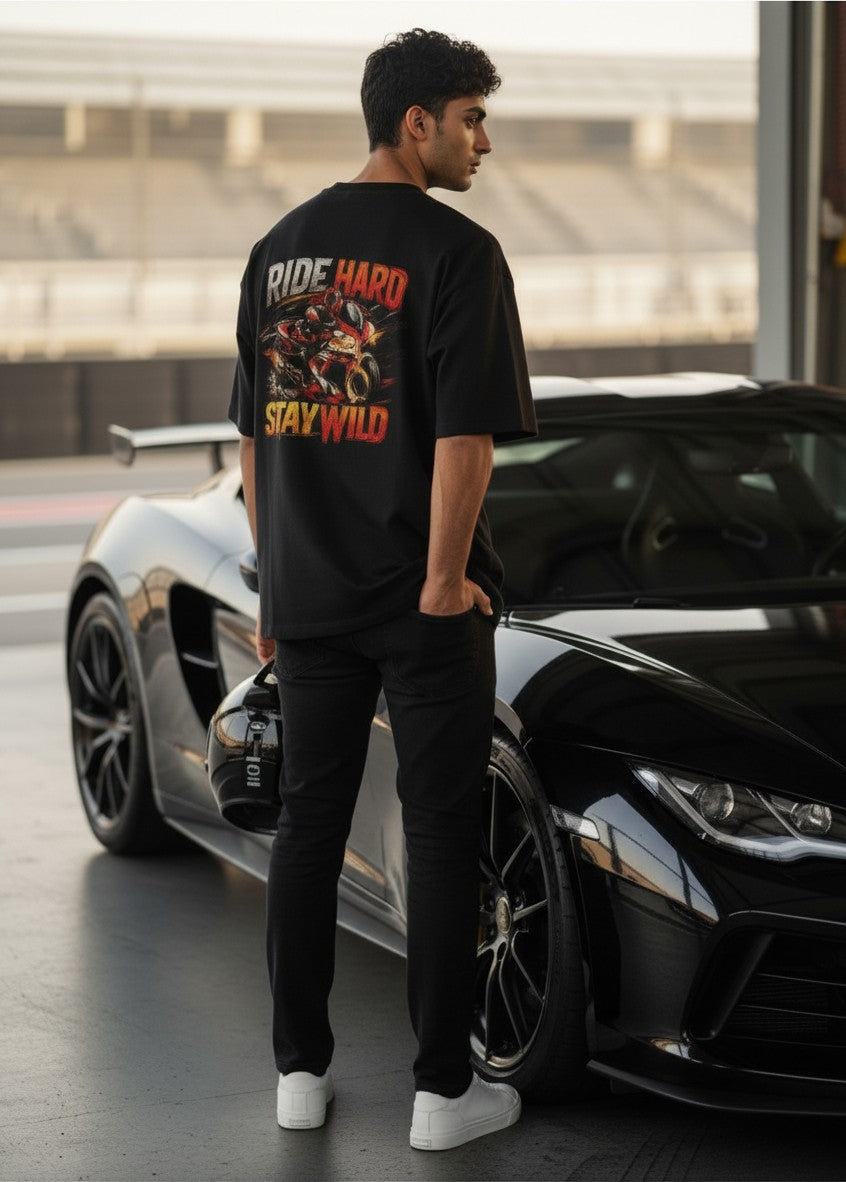 Print 999 oversized black cotton t-shirt with Ride Hard Stay Wild motorcycle graphic worn by a man at a racetrack.