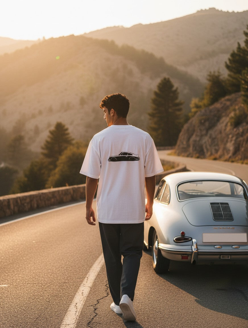 Man wearing Print 999 white oversized t-shirt with vintage car graphic on back, walking on a scenic mountain road.