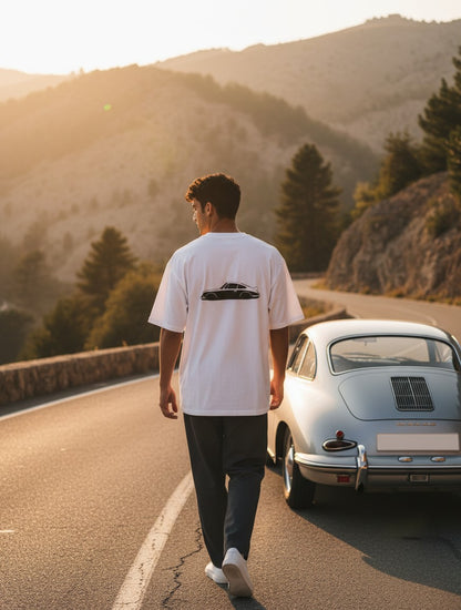 Man wearing Print 999 white oversized t-shirt with vintage car graphic on back, walking on a scenic mountain road.